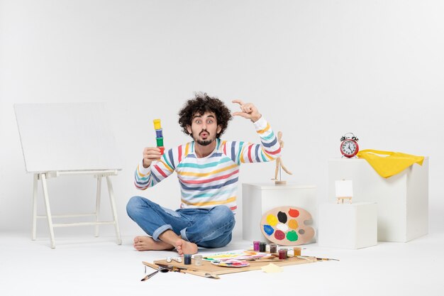 Vista frontal del macho joven sosteniendo pinturas para dibujar dentro de latas pequeñas en la pared blanca