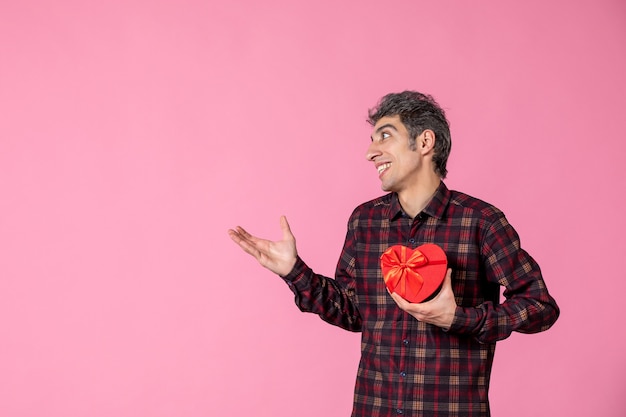 Vista frontal macho joven sosteniendo presente en forma de corazón rojo en la pared rosa