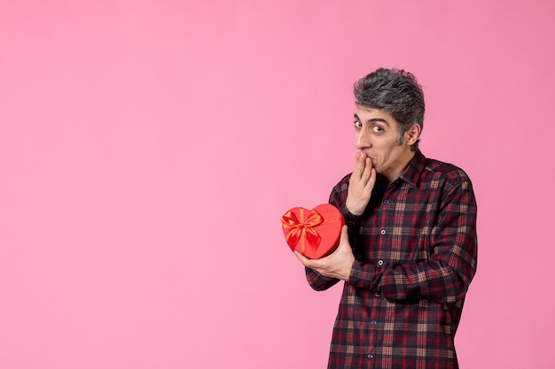 Vista frontal macho joven sosteniendo presente en forma de corazón rojo en la pared rosa