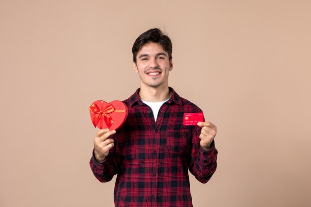 Vista frontal macho joven sosteniendo presente en forma de corazón y tarjeta bancaria en la pared marrón