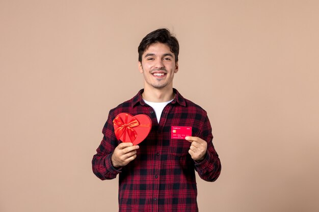 Vista frontal macho joven sosteniendo presente en forma de corazón y tarjeta bancaria en la pared marrón