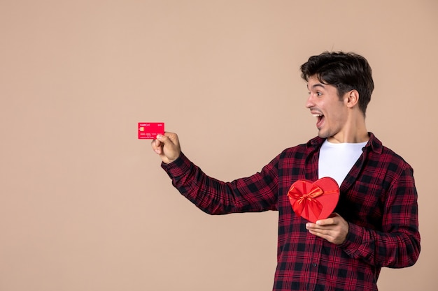 Vista frontal macho joven sosteniendo presente en forma de corazón y tarjeta bancaria en la pared marrón