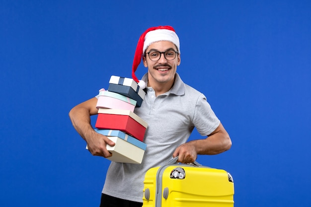 Vista frontal macho joven sosteniendo regalos y bolsa sobre fondo azul vacaciones de vuelos de avión