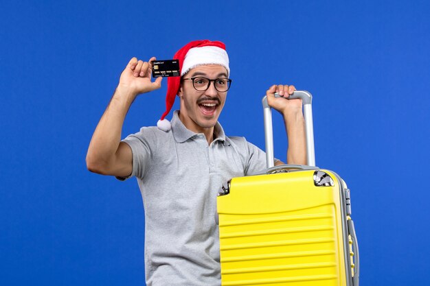 Vista frontal macho joven sosteniendo una tarjeta bancaria bolsa amarilla en la pared azul vuelos de aviones de vacaciones