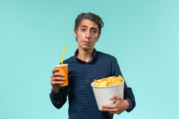 Vista frontal macho de mediana edad sosteniendo cips de patata y bebida sobre una superficie azul