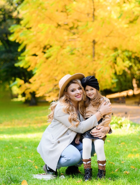 Vista frontal de una madre alegre con una hija encantadora vestida con ropa informal y abrazando sombreros y sentadas juntas entre el césped en el fondo