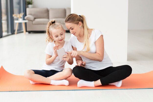 Vista frontal de la madre y la hija en casa en la estera de yoga jugando en el teléfono inteligente