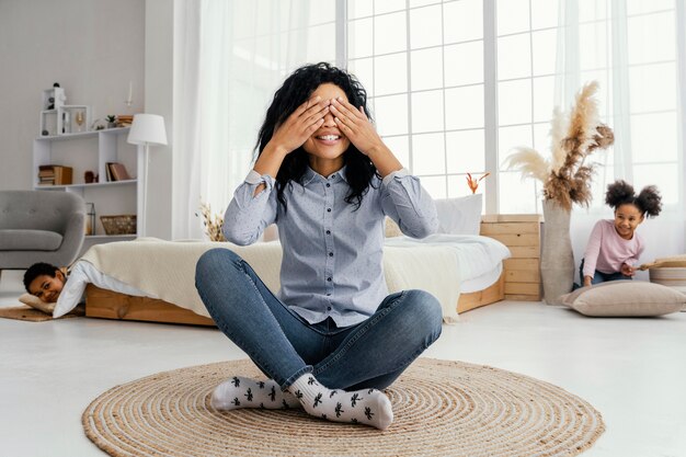 Vista frontal de la madre sonriente jugando al escondite en casa con sus hijos