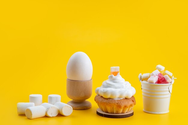 Una vista frontal de malvaviscos blancos junto con pasteles y caramelos en el escritorio amarillo, caramelo de azúcar color galleta dulce