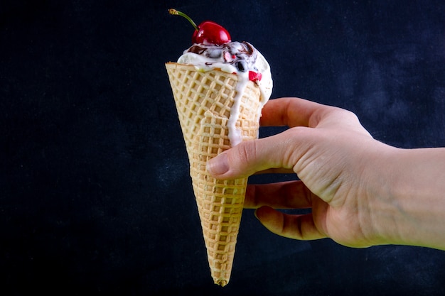 Vista frontal mano femenina sostiene helado con cereza en un cono de waffle en negro