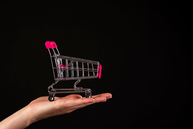 Vista frontal de una mano sosteniendo un mini carrito de compras sobre un fondo negro