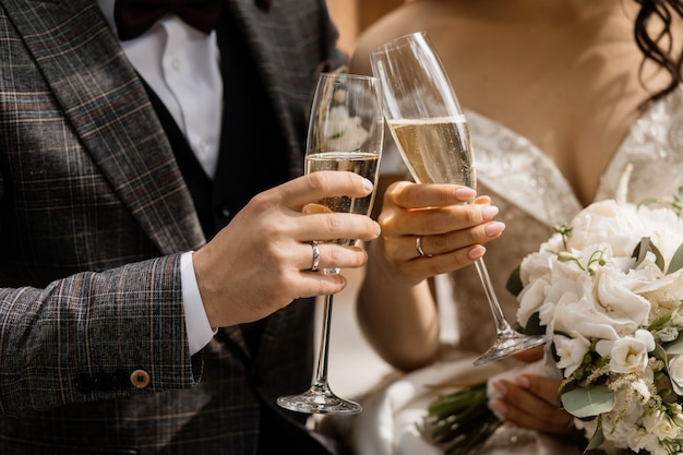 Vista frontal de las manos de los novios con copas de champán y bouquet de bodas