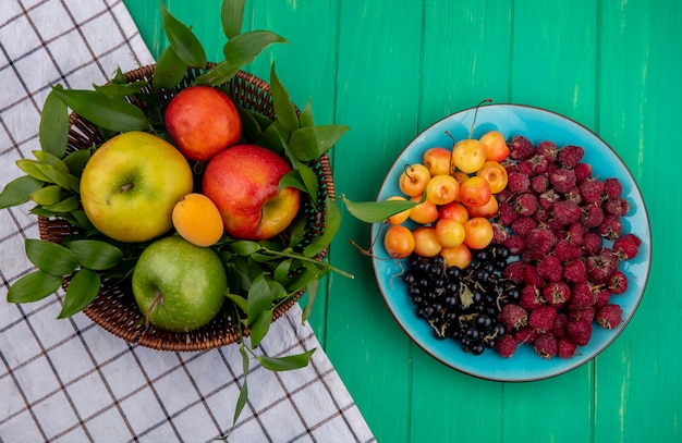 Vista frontal de manzanas de colores en una canasta con cerezas blancas, frambuesas y grosellas negras en una placa sobre una superficie verde