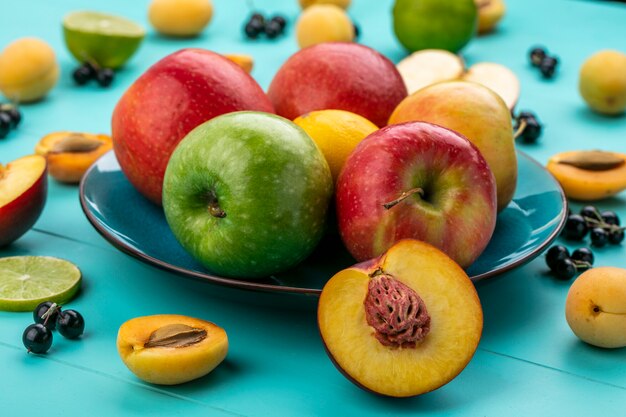 Vista frontal de manzanas de colores en un plato con albaricoques cal y grosellas negras sobre una superficie azul claro