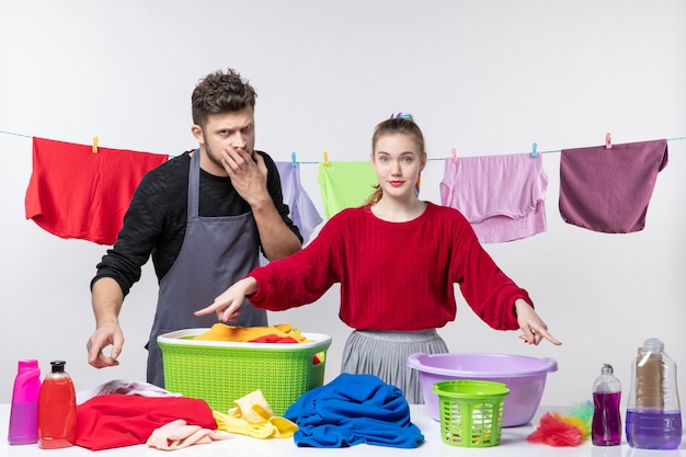 Vista frontal del marido de limpieza y su esposa apuntando a detergentes canasta de lavandería y productos de limpieza en la mesa en la pared blanca