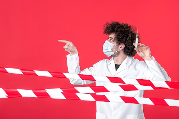 Vista frontal médico masculino con inyección contra virus sobre fondo rojo advertencia covid- uniforme de cuarentena médica de salud