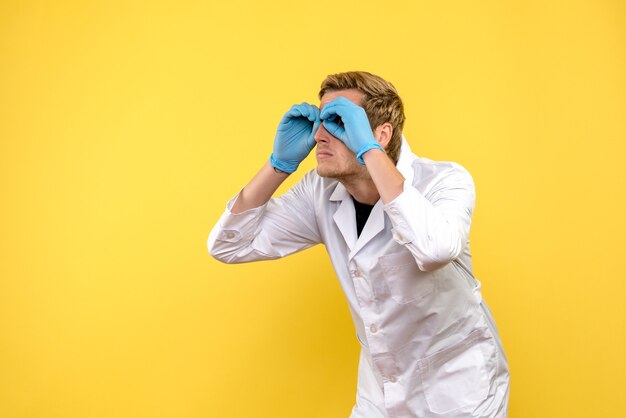 Vista frontal médico masculino mirando a través de los dedos sobre fondo amarillo covid- salud pandémica