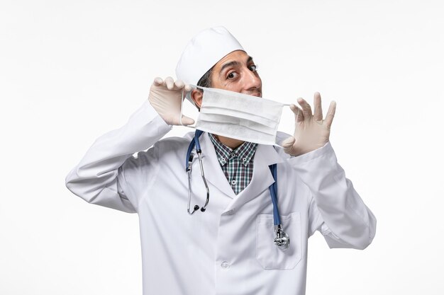 Vista frontal médico masculino en traje médico blanco debido al coronavirus con máscara en el escritorio blanco