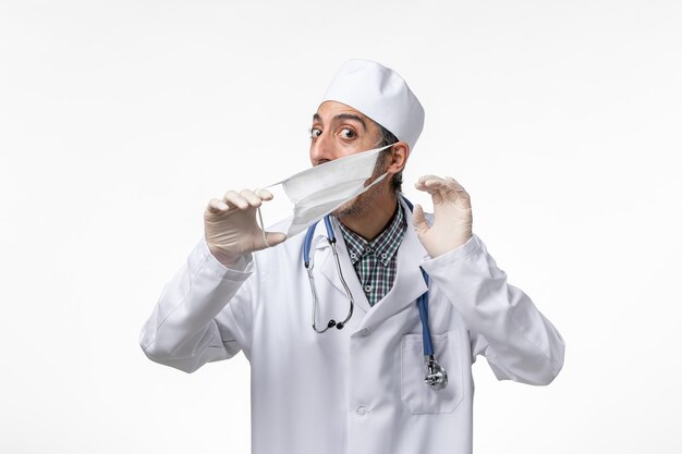 Vista frontal médico masculino en traje médico blanco debido al coronavirus con máscara en una superficie blanca clara