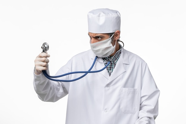 Vista frontal médico masculino con traje médico blanco con máscara debido al coronavirus con estetoscopio azul sobre fondo blanco pandemia de virus de enfermedad hospitalaria covid