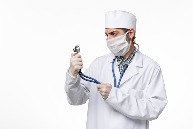 Vista frontal médico masculino con traje médico blanco y máscara debido al coronavirus con estetoscopio en el escritorio blanco