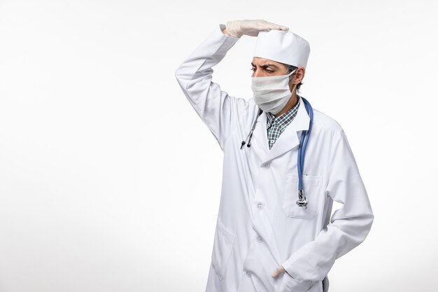 Vista frontal médico masculino en traje médico blanco con máscara debido al coronavirus posando sobre una superficie blanca clara