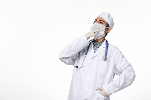 Vista frontal médico masculino con traje médico blanco y máscara debido al coronavirus en la superficie blanca