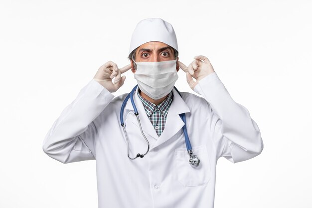 Vista frontal médico masculino en traje médico blanco con máscara debido al coronavirus en la superficie blanca