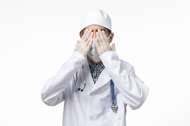 Vista frontal médico masculino en traje médico blanco con máscara debido a covid- sobre superficie blanca
