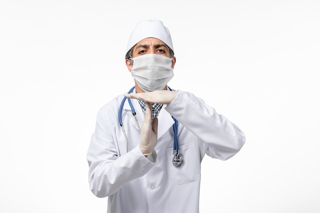 Vista frontal médico masculino en traje médico blanco con máscara debido a covid en superficie blanca clara