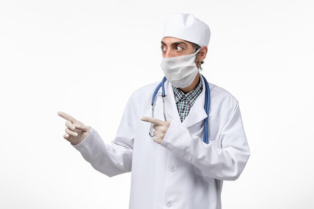 Vista frontal médico masculino en traje médico blanco con máscara debido a covid en superficie blanca