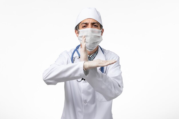Vista frontal médico masculino en traje médico blanco con máscara debido a covid en superficie blanca