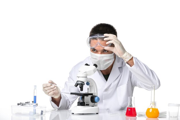 Vista frontal médico masculino en traje médico blanco con máscara debido a covid usando microscopio en el espacio en blanco