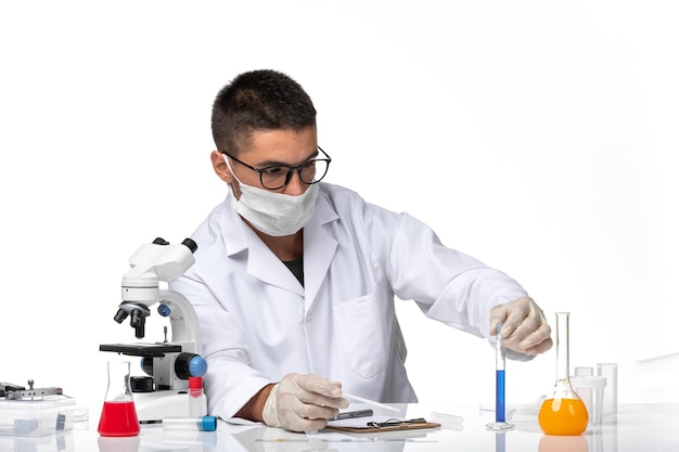 Vista frontal médico masculino en traje médico blanco y con máscara que trabaja con una solución en un espacio en blanco claro