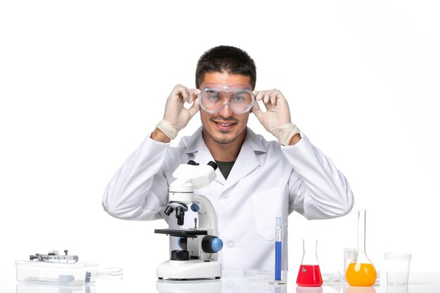 Vista frontal médico masculino en traje médico blanco sentado con soluciones sonriendo en el espacio en blanco