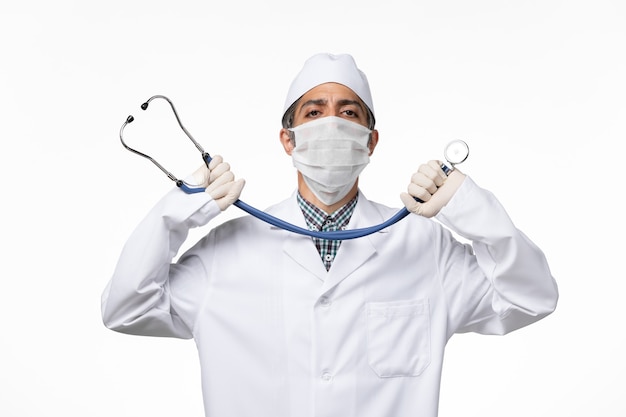 Vista frontal médico masculino con traje médico estéril y máscara debido al coronavirus con estetoscopio sobre una superficie blanca
