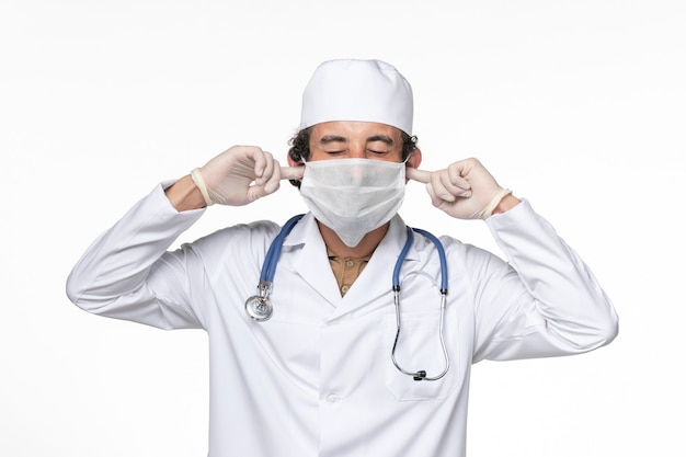 Vista frontal médico masculino en traje médico con máscara como protección contra el covid: cerrar los oídos en la pared blanca medicina pandémica del virus del coronavirus