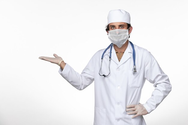 Vista frontal médico masculino en traje médico con máscara como protección contra el covid- en la enfermedad del virus de escritorio blanco covid- pandemia