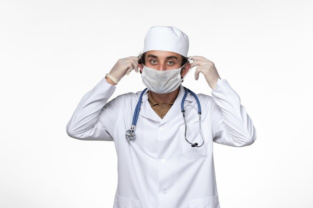 Vista frontal médico masculino en traje médico con máscara como protección contra el covid- en el escritorio blanco enfermedad por virus covid-pandemia de salud