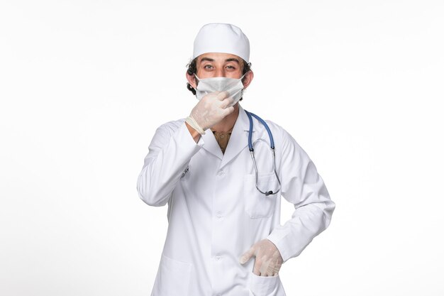 Vista frontal médico masculino en traje médico con máscara como protección contra covid- en la medicina pandémica del virus coronavirus de pared blanca clara