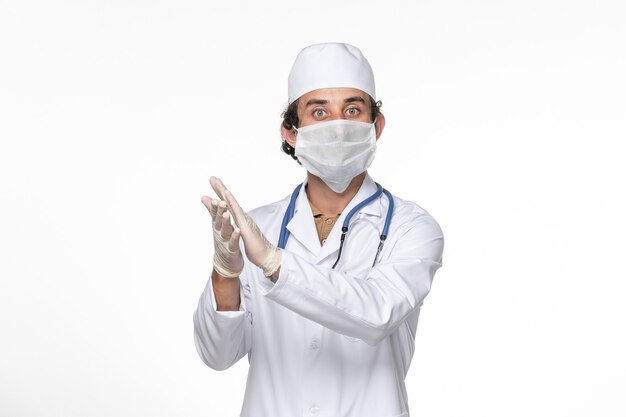 Vista frontal médico masculino en traje médico con máscara como protección contra el covid en la medicina pandémica del virus del coronavirus de la pared blanca