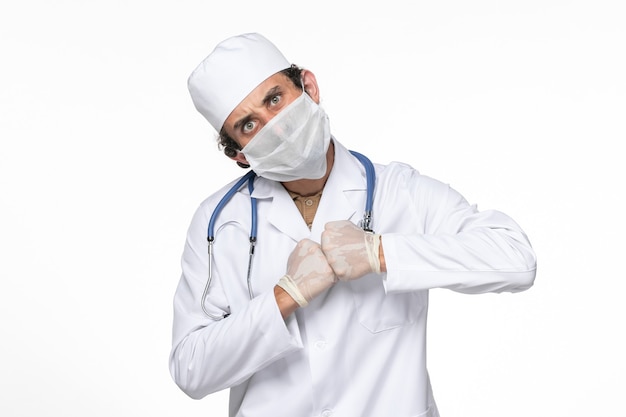 Vista frontal médico masculino en traje médico con máscara como protección contra el covid en la pared blanca.