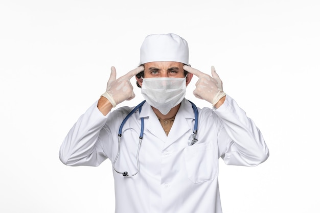 Vista frontal médico masculino en traje médico con máscara como protección contra el covid: pensamiento en la pared blanca, salpicadura de virus, coronavirus, pandemia