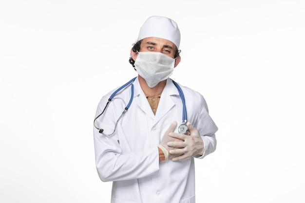 Vista frontal médico masculino en traje médico con máscara como protección contra el covid: tener dolor de corazón en la pared blanca.