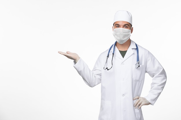 Vista frontal médico masculino en traje médico y con una máscara contra el covid con estetoscopio en la pared blanca virus covid- enfermedad pandemia