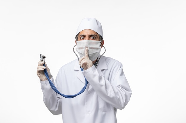 Vista frontal médico masculino en traje médico y máscara debido al coronavirus con estetoscopio sobre superficie blanca