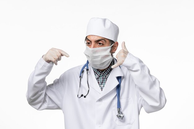 Vista frontal médico masculino en traje médico con máscara debido a covid- en el escritorio blanco