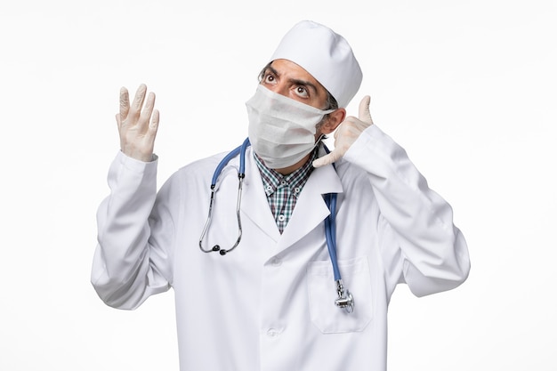 Vista frontal médico masculino en traje médico con máscara debido a covid en superficie blanca