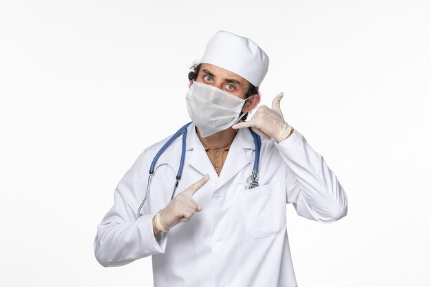 Vista frontal médico masculino en traje médico con máscara estéril como protección contra el covid en la enfermedad pandémica del coronavirus del virus de la pared blanca