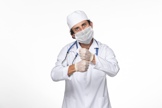 Vista frontal médico masculino en traje médico con máscara estéril como protección contra el covid en la pandemia de coronavirus de virus de salpicaduras de pared blanca